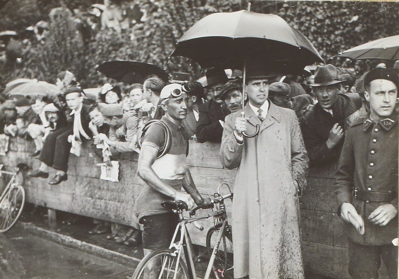 Photographie sépia : un coureur cycliste, lunettes relevées sur le casque et maillot mouillé, tient son vélo à l'arrivée d'une étape. À côté de lui, un homme en imperméable taché de boue tient un grand parapluie ; derrière une palissade de bois, une foule de spectateurs, plusieurs parapluies ouverts.