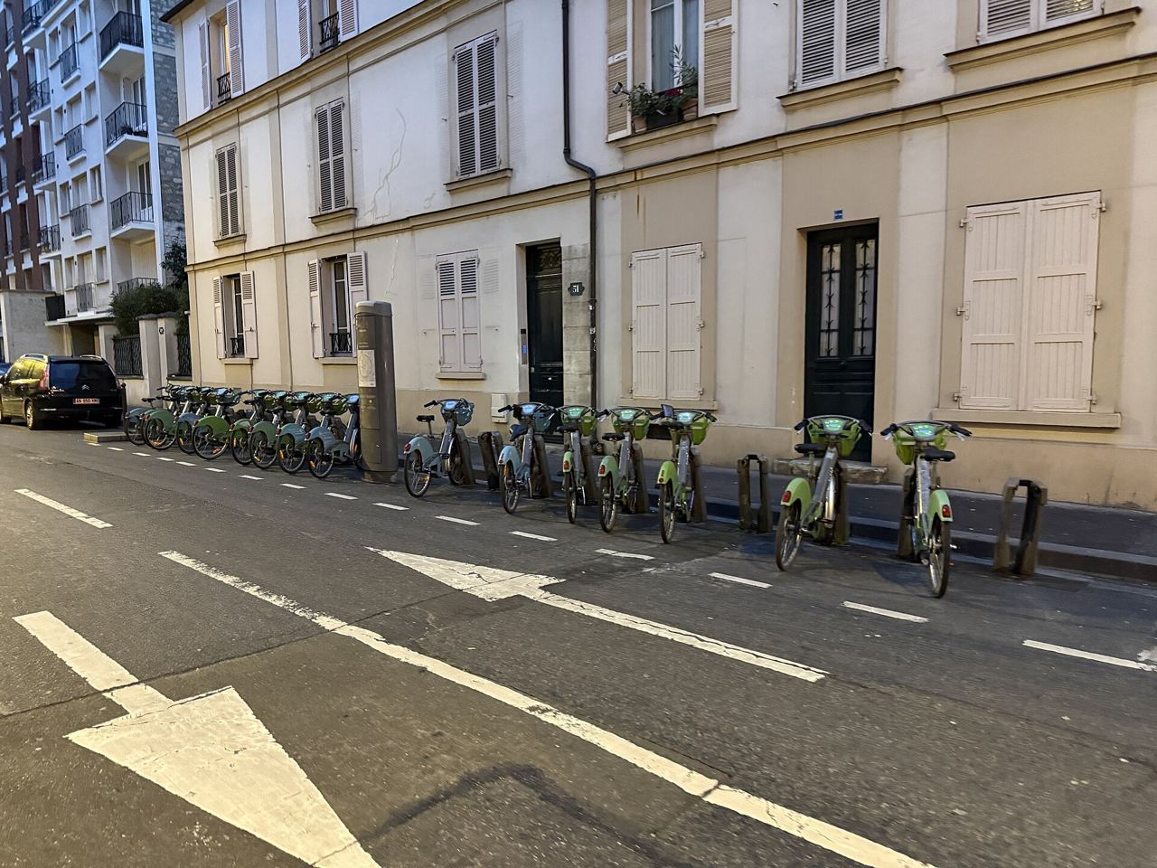 Station Vélib' Fessart – Château à Boulogne-Billancourt : des vélos en libre-service verts et bleus alignés le long d'une façade crème, sur une rue aux marquages blancs.