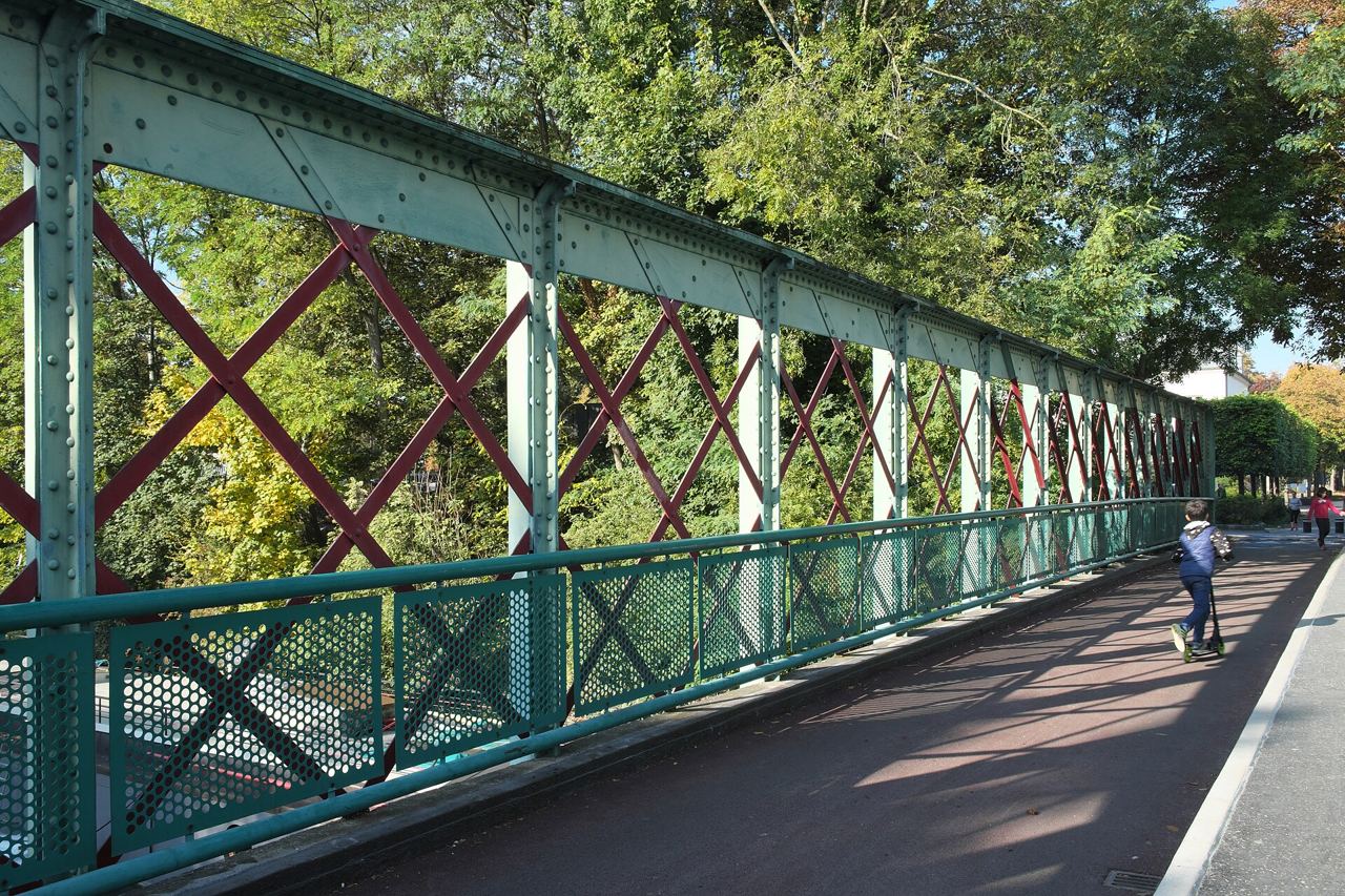 La passerelle métallique verte et rouge de l'île Saint-Germain à Issy-les-Moulineaux, avec son tablier au revêtement rouge sur lequel passe une personne en trottinette, entre les arbres.